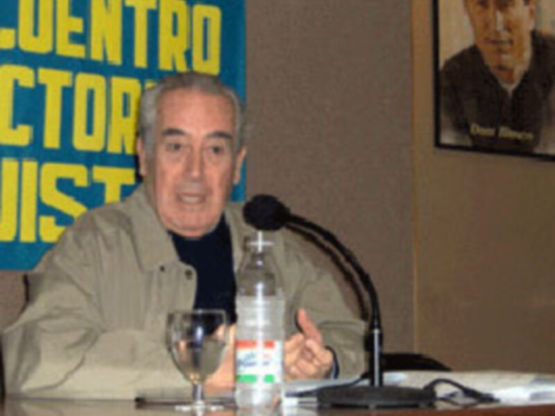 Persona hablando en un evento público, con un cartel de 'Encuentro Elector' en el fondo y un vaso de agua sobre la mesa.