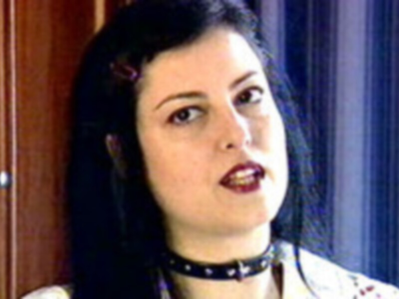 Una persona con cabello largo y oscuro, llevando una gargantilla de cuero negra y una camisa blanca con detalles de color rojo.