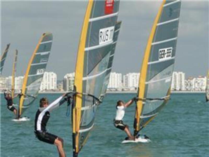 Competición de vela de viento en el mar con barcos a vela y edificios en el horizonte.
