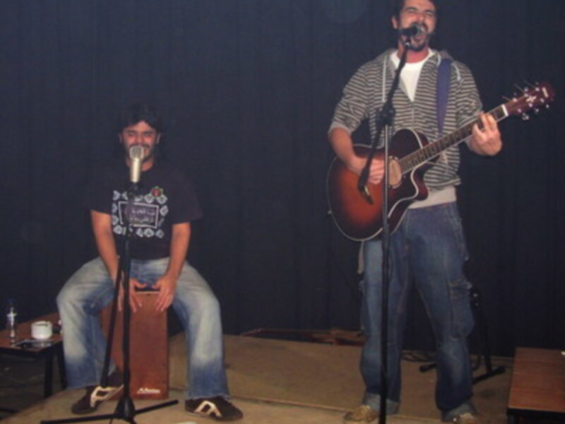 Dos personas en un escenario, una tocando guitarra y la otra cantando en micrófono. Ambas están vestidas con camisetas y pantalones de mezclilla. El fondo es un telón negro.