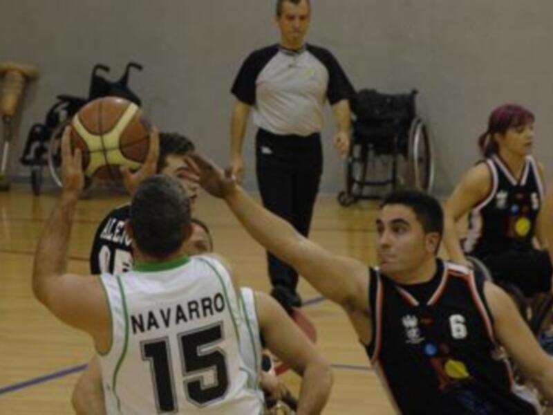 Jugadores de baloncesto en una acción de juego, con un árbitro en el centro y un ambiente deportivo en el fondo.
