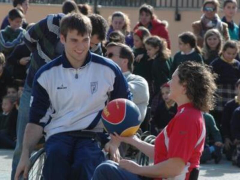 Joven en silla de ruedas jugando baloncesto con un niño en el suelo, rodeados por un grupo de espectadores.