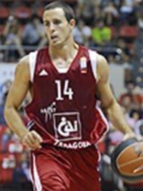 Un jugador de baloncesto con el número 14 en su camiseta roja, posando con una pelota de baloncesto en la mano. El fondo muestra una tribuna de espectadores y otros jugadores en el estadio.