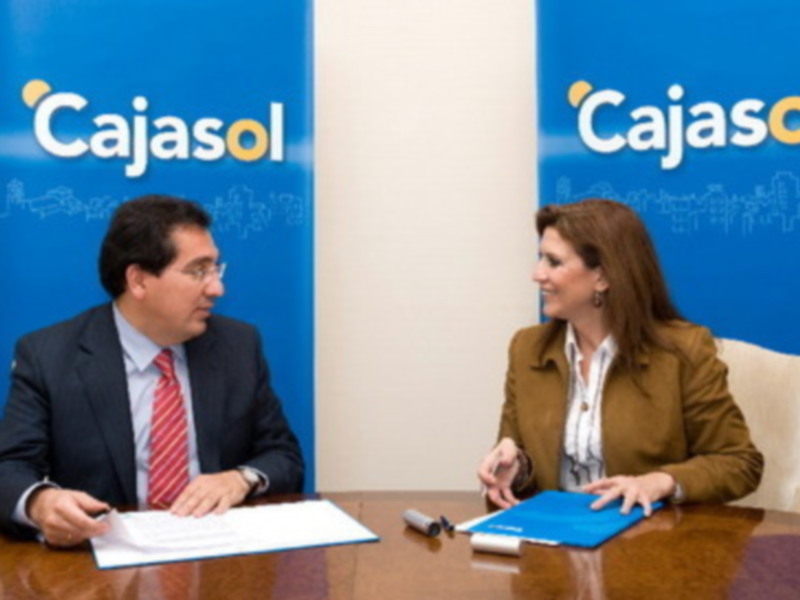 Dos personas sentadas en una mesa, uno de ellos con una camisa blanca y corbata roja, el otro con una chaqueta marrón. En el fondo, dos carteles con el logotipo de Cajasol.