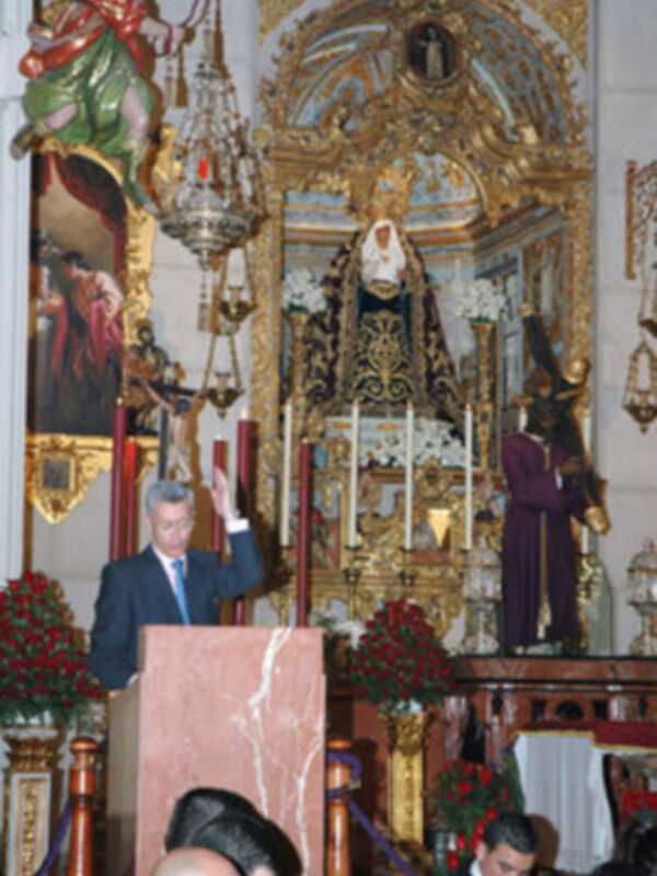 Discurso en un evento religioso frente a un altar ornamental con imágenes religiosas y personas en trajes formales.