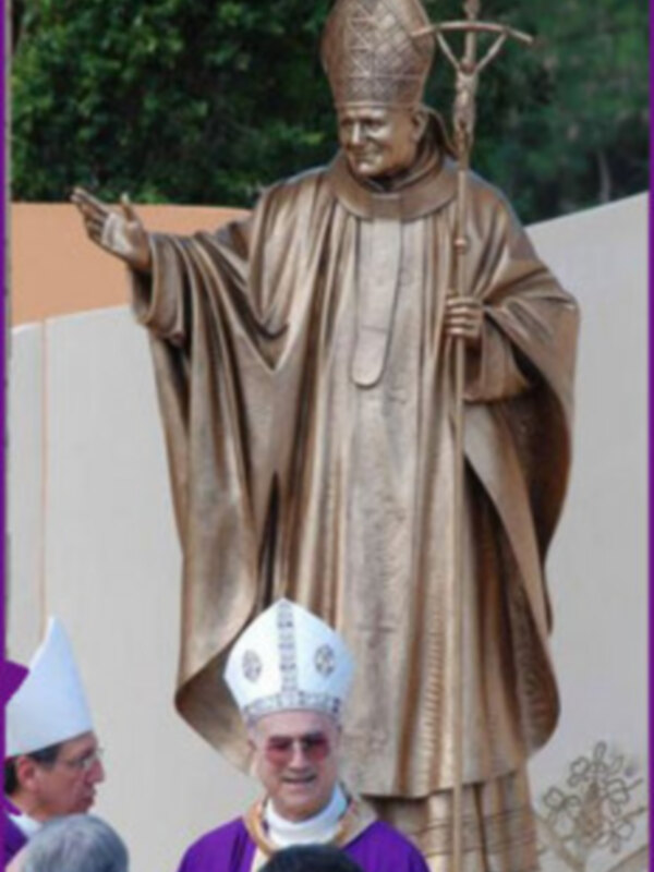 Estatua de bronce de un sacerdote con hábito y mitra, rodeada por personas en hábitos sacerdotales.