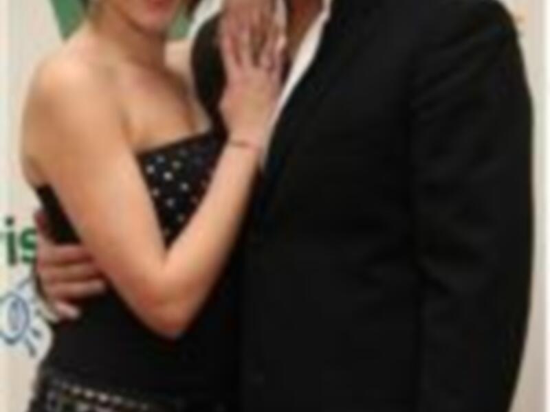 Una pareja sonríe en un evento formal, ella con un vestido negro y él con un traje negro, sosteniendo un ramo de flores.