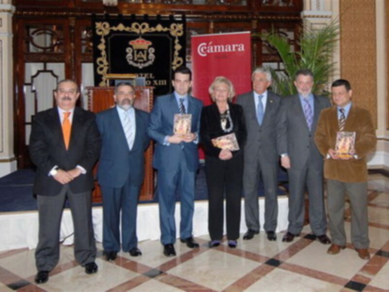 Una imagen de un grupo de personas en un evento formal, posando con una persona que sostiene un libro. En el fondo, se puede ver una bandera con el nombre 'Cámara' y un escudo de armas. El suelo es de mármol con patrones geométricos.
