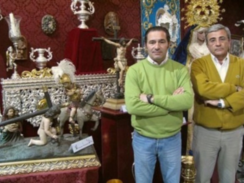 Dos hombres en una sala decorada con objetos religiosos y artísticos, incluyendo esculturas y altares.