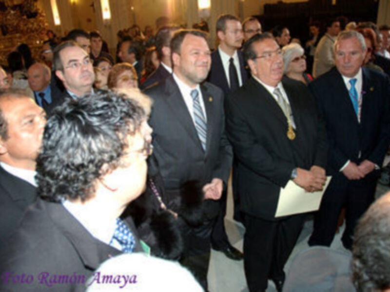 Conferencia o evento formal en un interior iluminado, con personas vestidas de traje y semblante serio.