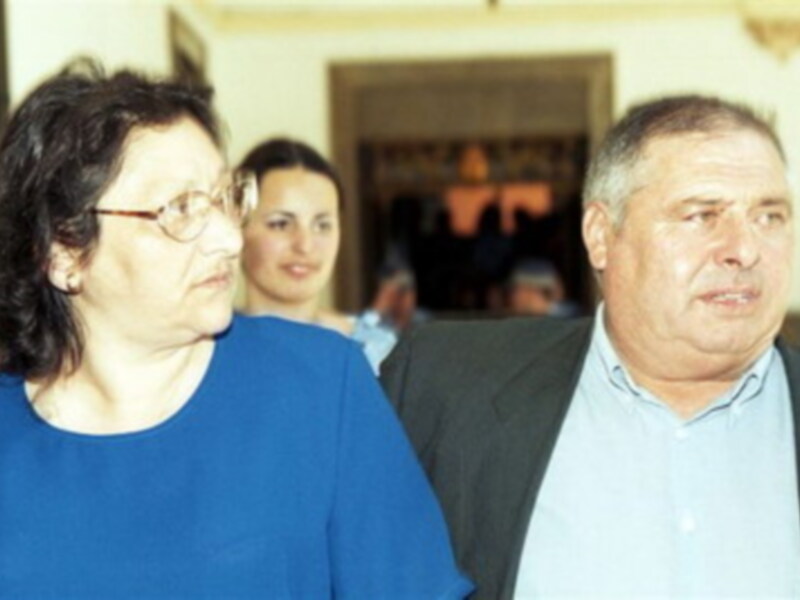 Una mujer con gafas y una mujer con una camisa azul, ambos están mirando hacia la cámara. Detrás de ellos, hay personas que parecen estar en una reunión o evento. La imagen parece ser de una reunión formal o un evento público.