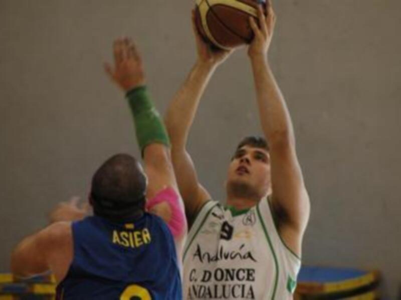 Jugador encestando mientras un defensor intenta bloquear durante un partido de baloncesto en Andalucía.