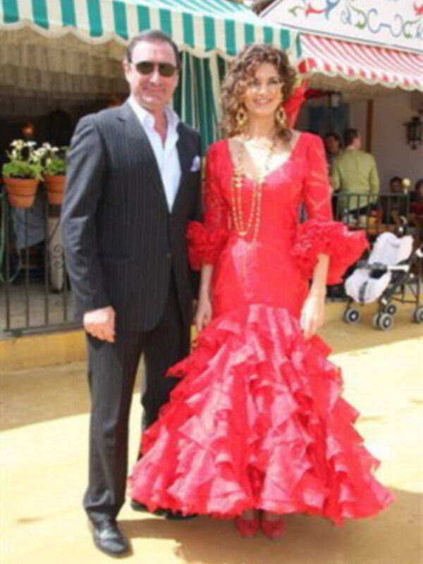 Una pareja está posando para una foto. La mujer lleva un vestido rojo con volantes y una corona de flores en la cabeza. El hombre está vestido con un traje negro y gafas de sol. En el fondo, se pueden ver personas y una carpa con un letrero que dice 'S'.