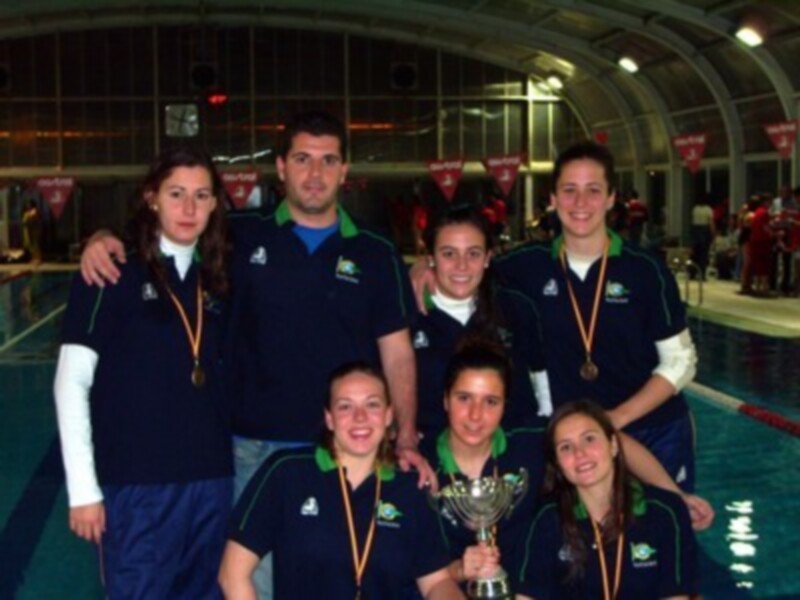Equipo de natación celebrando con trofeo y medallas en piscina cubierta.