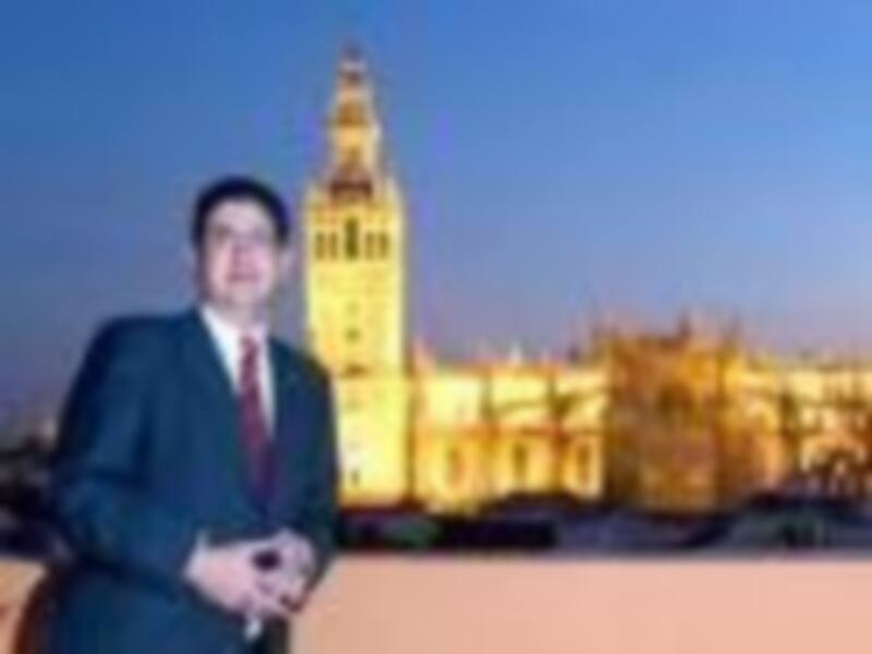 Un hombre en traje formal con una corbata azul frente a la catedral de Seville en la noche, iluminada por luces de color amarillo y rojo.