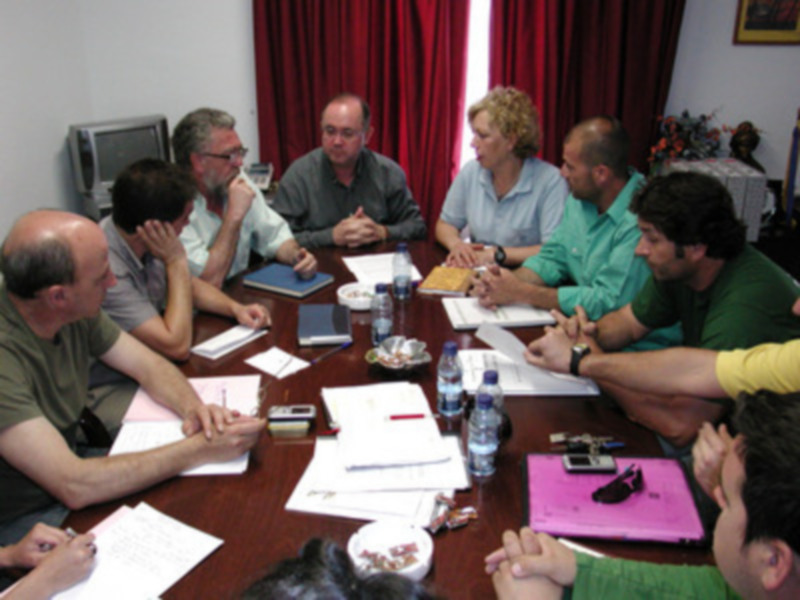 Reunión de trabajo en una mesa redonda con documentos y agua en el centro.