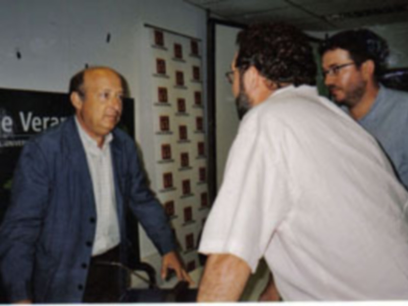 Tres personas conversando en un ambiente interior con una pared de cuadros en el fondo.