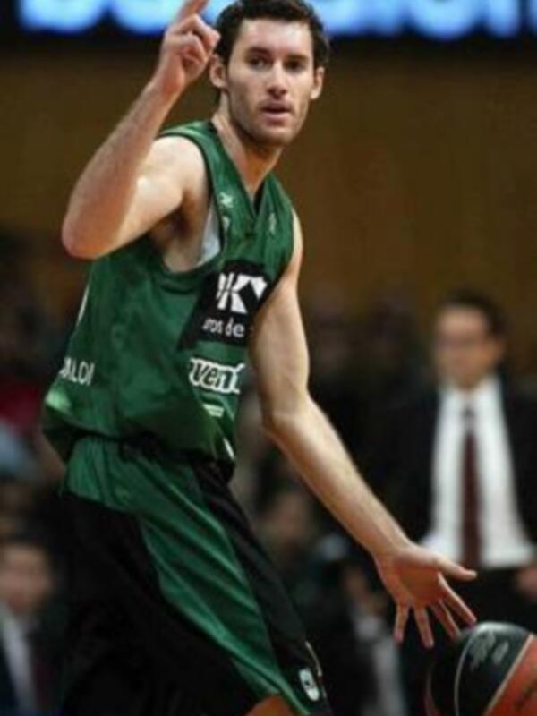 Un jugador de baloncesto en acción, con una camiseta verde y negra, sosteniendo un balón y levantando el dedo índice.