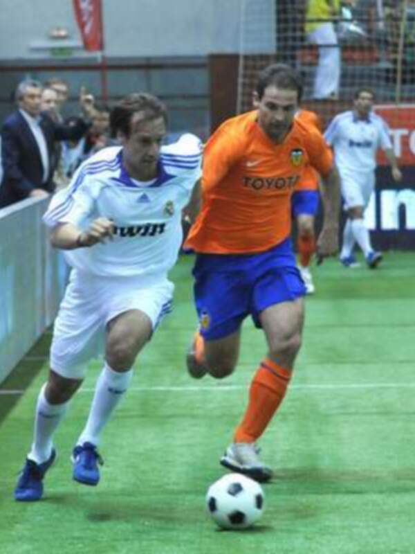 Dos jugadores de fútbol en acción, uno con uniforme blanco y otro con uniforme naranja y azul, en un campo de juego con una publicidad de Toyota en el fondo.