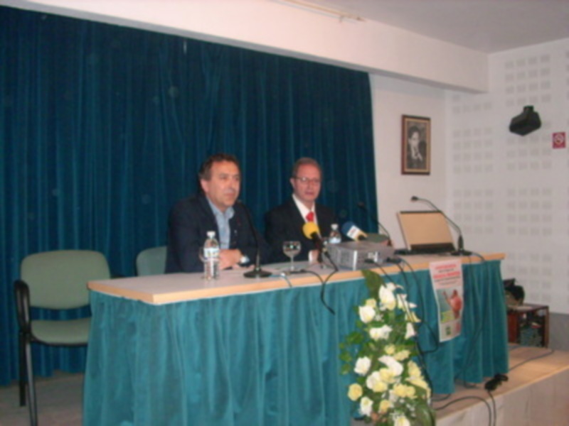 Dos hombres en trajes sentados detrás de una mesa con micrófonos y un botón de agua. La mesa está cubierta con una falda azul, hay flores blancas y un cartel informativo. La pared de fondo tiene cortinas verdes y una foto colgada.