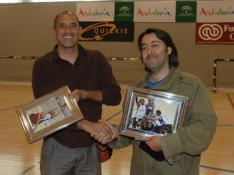 Dos personas sonrientes sostienen marcos de fotos, uno con un equipo de fútbol y el otro con un equipo de baloncesto, en un contexto deportivo.