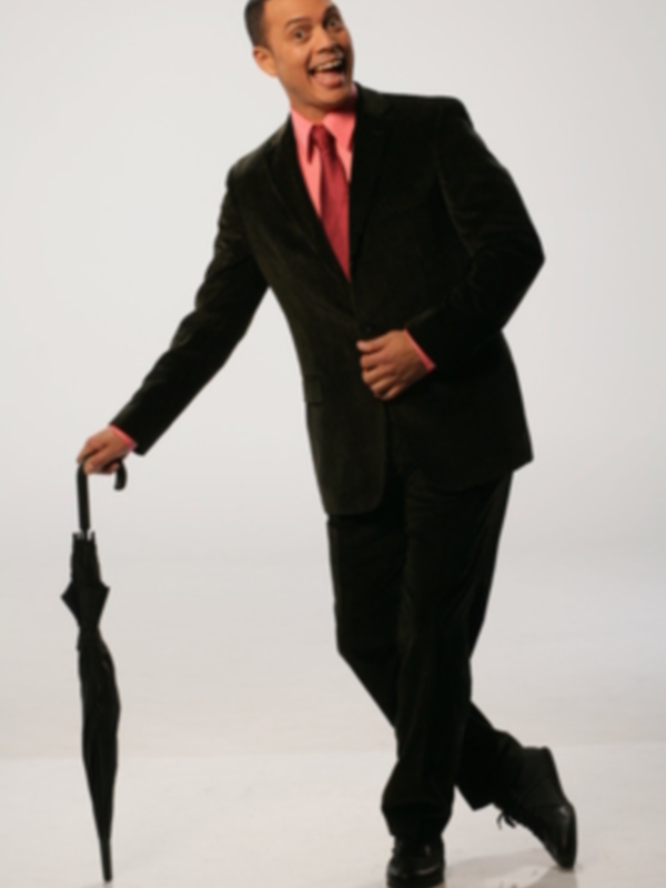 Un hombre en traje negro y corbata rosa, caminando con un paraguas negro en la mano izquierda, contra un fondo blanco.