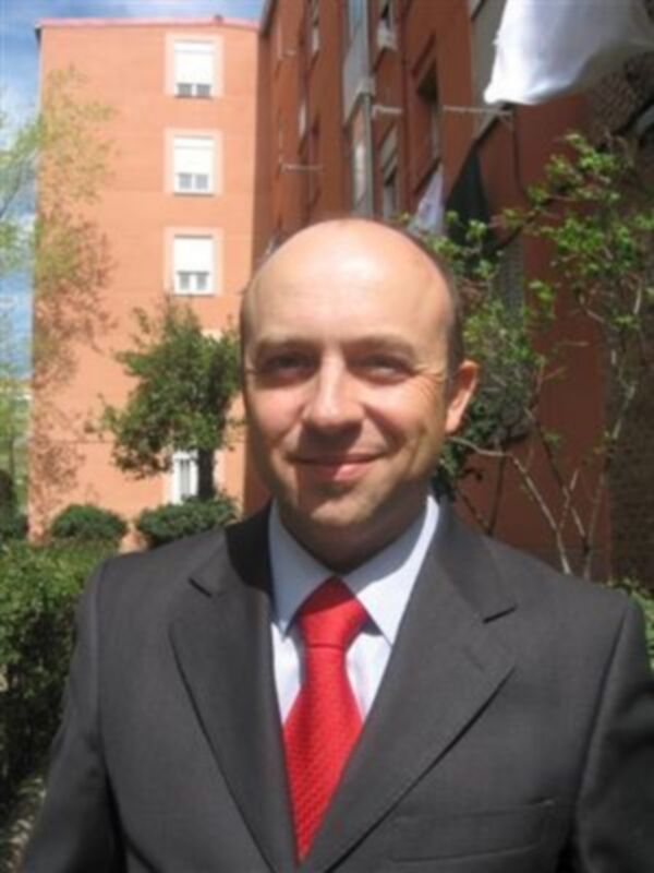 Hombre en traje negro con corbata roja, sonriendo frente a un edificio rojo y verde.