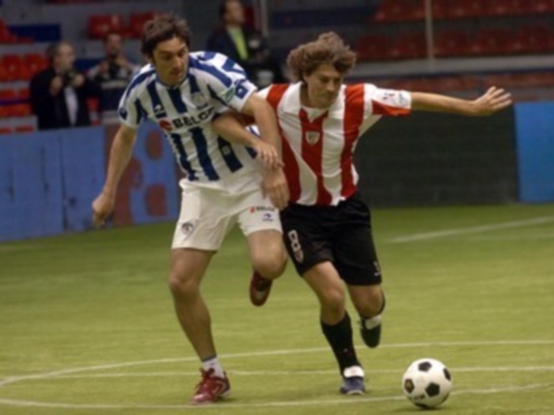 Dos jugadores de fútbol en acción, uno con camiseta azul y blanco y el otro con camiseta roja y blanco, luchando por controlar el balón en un campo de juego.