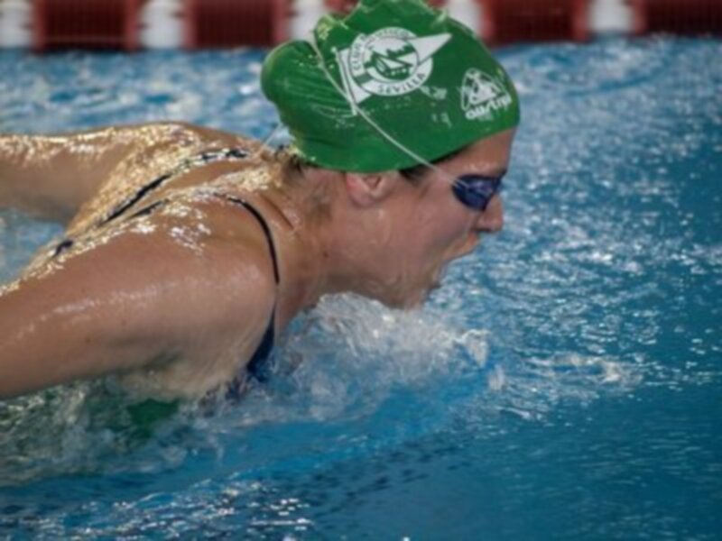 Una nadadora en acción, con una gorra verde y un traje de baño negro, en una piscina de agua azul claro.