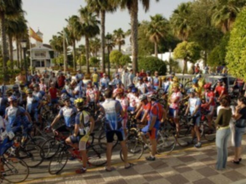 Ciclistas y espectadores se reúnen en una calle con palmeras y edificios históricos en el fondo, antes de una carrera de ciclismo.