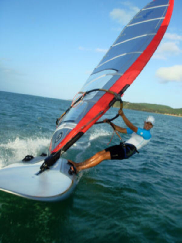 Un windsurfer realiza una maniobra en el agua azul profundo, con un cielo azul claro y una isla en el fondo.