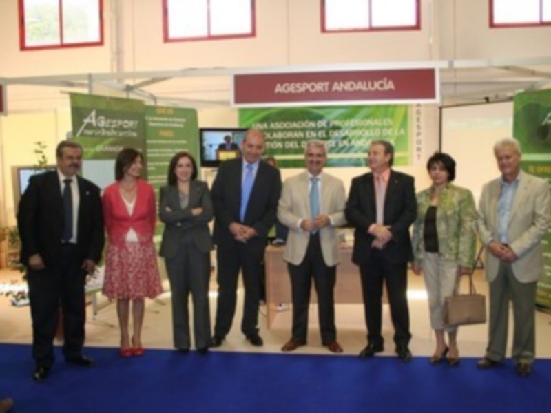 Una imagen de un evento empresarial en Andalucía, con varios empresarios y empresarias posando en frente de un stand de la entidad AGEPSORT ANDALUCÍA. Los stands están decorados con banderas y carteles informativos.
