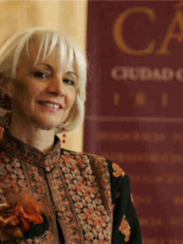 Una mujer con cabello blanco y una chaqueta estampada, posando frente a un fondo rojo con texto en español.