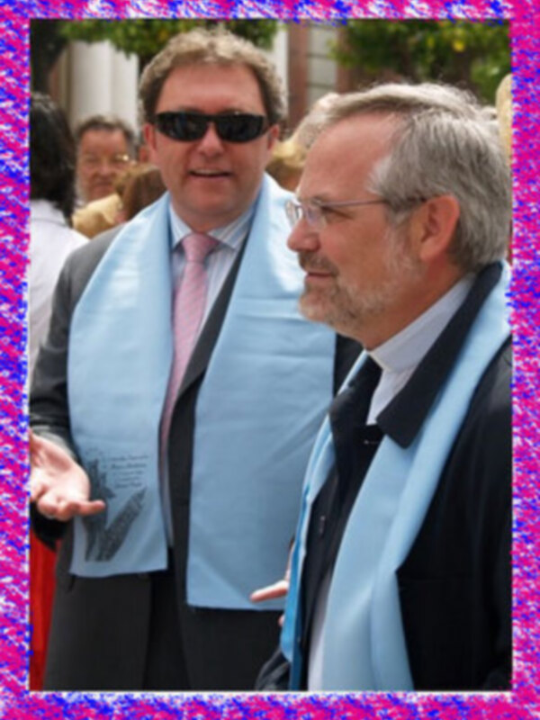 Dos hombres en traje conversando al aire libre, uno con gafas de sol y el otro con una camisa de manga larga y corbata.