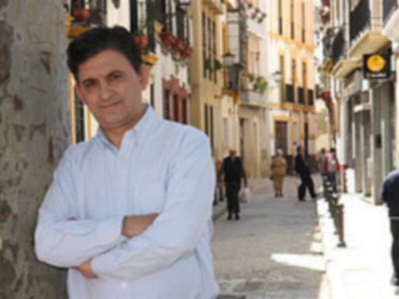 Un hombre en una calle de la ciudad, posando con las manos cruzadas en el pecho, mientras otros ciudadanos caminan al fondo.