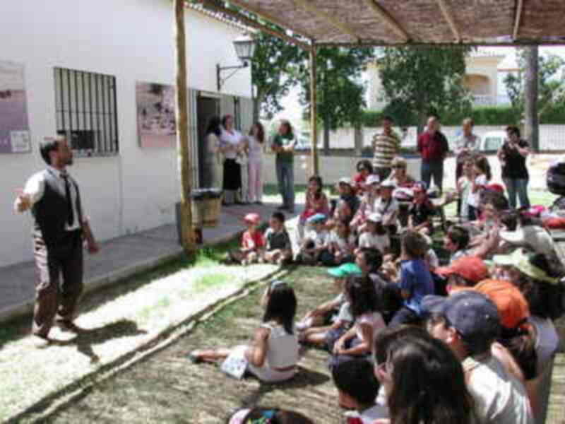 Un grupo de niños y adultos se reúnen al aire libre en un patio, con una persona en el centro del grupo. La escena parece ser una reunión o una actividad educativa.