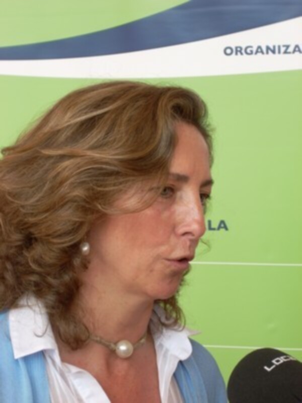 Una mujer con cabello largo y ondulado, usando una camisa blanca y un collar de perlas, hablando en un micrófono. El fondo es verde con un logotipo de 'ORGANIZACIÓN' en el cielo azul.