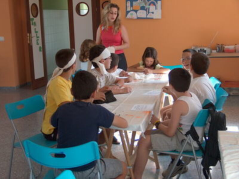 Niños sentados en una mesa en un aula, con una maestra en el centro.