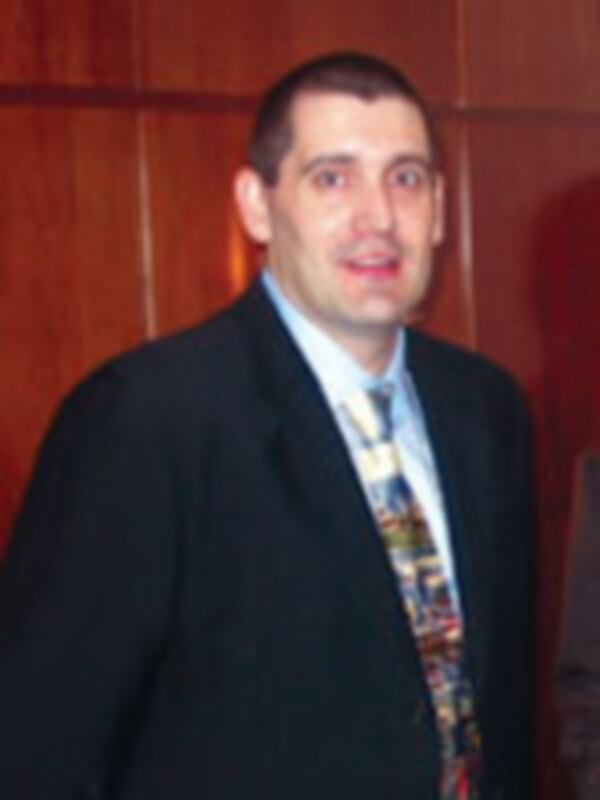 Un hombre en traje y corbata está sonriendo en una imagen de interior con un fondo de madera.
