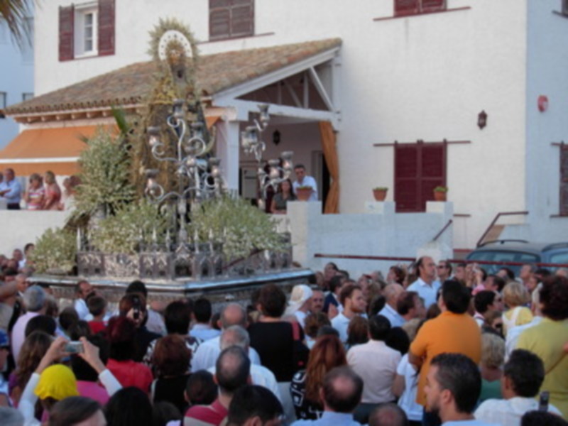 Una procesión religiosa con una imagen de la Virgen en el centro, rodeada por una multitud de fieles. La imagen se encuentra en una calle con edificios blancos y ventanas con persianas.