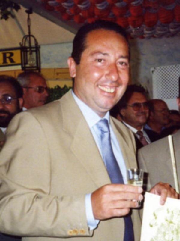 Hombre sonriente en traje con camisa azul y corbata, sosteniendo un vaso de champán en un evento social.