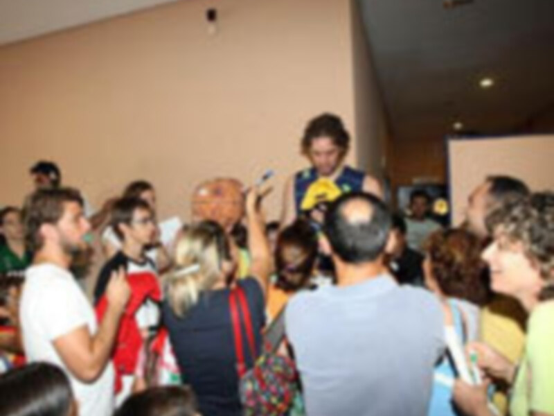 Una escena de una fiesta donde una mujer con un vestido azul y un ramo de flores se dirige hacia la cámara, rodeada de personas que parecen estar disfrutando del evento.