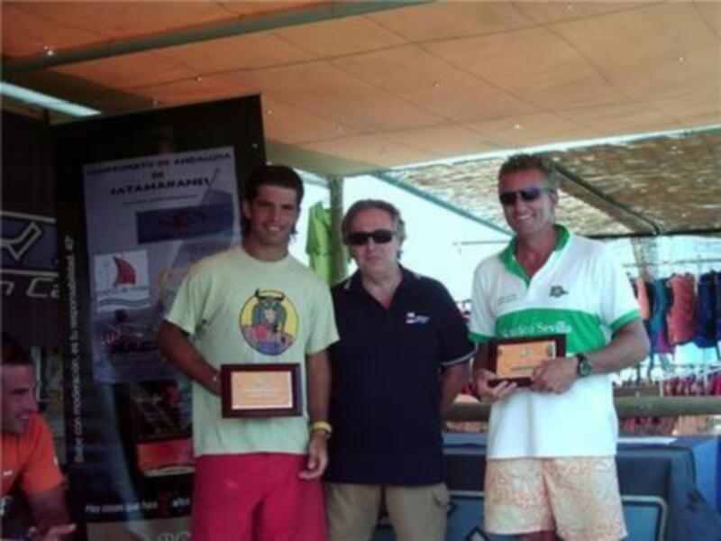 Tres personas posan con trofeos en un evento deportivo bajo una estructura sombreada.