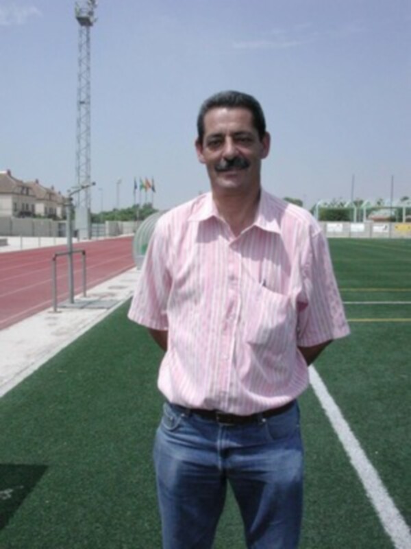 Un hombre en una camisa rosa y pantalones azules, posando en un estadio de atletismo con una bandera en el fondo.