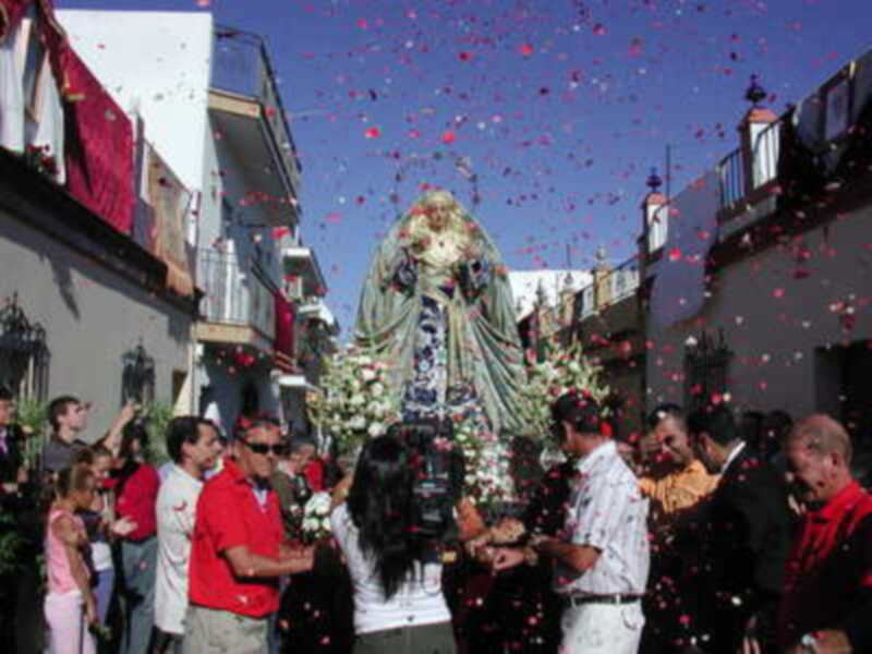 Fiesta religiosa con la procesión de una imagen venerada, rodeada de personas y confeti en el cielo azul.