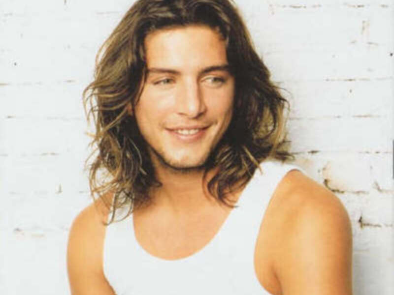 Un hombre con cabello largo y una sonrisa amable, posando frente a una pared de ladrillos blancos.