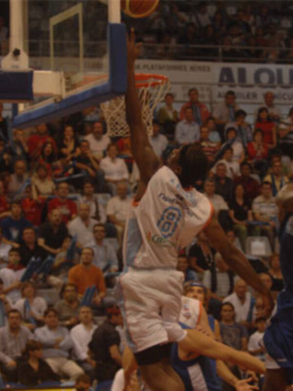 Jugador en acción durante un partido de baloncesto, saltando para hacer un mate en un entorno de público animado.