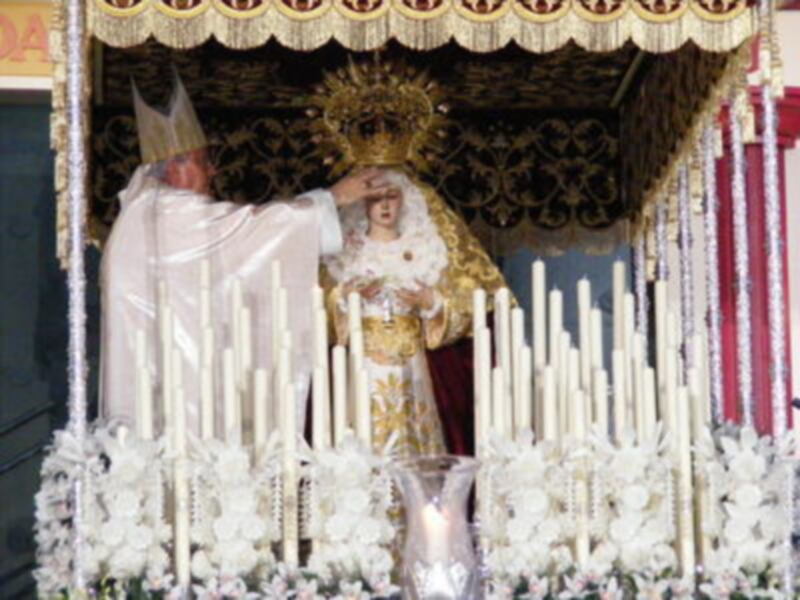 Estatua de la Virgen de la Candelaria en un altar con velas y un manto blanco, rodeada por un manto de flores y un velo dorado.