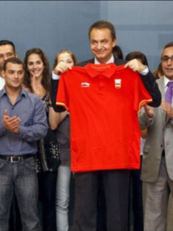 Persona sosteniendo una camiseta roja con logotipos, rodeada por un grupo de personas en una reunión formal.