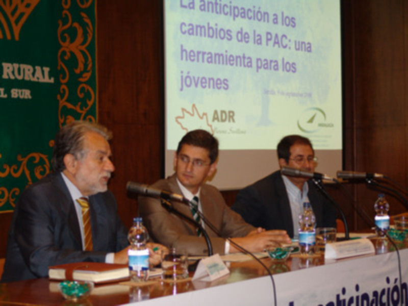 Tres hombres en trajes sentados en una mesa con micros y libros, con un fondo que incluye una pantalla con texto en español sobre la PAC y ADR.